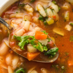 A close-up of a bowl of vegan minestrone soup with vegetables, pasta, and greens, with a spoon lifting a portion. Text reads "Slow Cooker Minestrone Soup." Perfect for your slow cooker!.