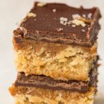 Two stacked Chocolate Caramel Bars with a crumbly base, a luscious middle layer, and a smooth chocolate topping, sprinkled with flakes of sea salt. These no-bake treats are perfect for anyone craving a delicious vegan pudding.