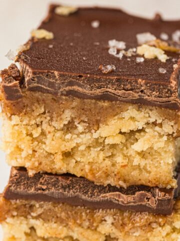 Two stacked Chocolate Caramel Bars with a crumbly base, a luscious middle layer, and a smooth chocolate topping, sprinkled with flakes of sea salt. These no-bake treats are perfect for anyone craving a delicious vegan pudding.