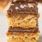 Three stacked no-bake vegan chocolate caramel bars topped with flaky salt, showcasing distinct chocolate, caramel, and biscuit-like layers. Text above reads "Vegan Chocolate Caramel Bars.