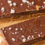 Vegan chocolate caramel bars topped with flaky sea salt, showing luscious layers of caramel and shortbread on baking parchment—an irresistible no-bake treat.
