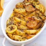 Oven-baked potato wedges in a white dish, topped with herbs and slices of roasted lemon—an easy Vegan Greek Lemon Potatoes recipe perfect for any garlic recipe lover.