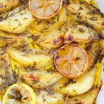 Close-up of easy Vegan Greek Lemon Potatoes, baked to perfection and garnished with herbs, garlic, and lemon slices in a white dish.