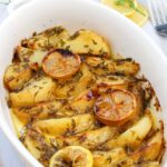 A white dish filled with easy Vegan Greek Lemon Potatoes, garnished with lemon slices and herbs, sits on a table with a fork and lemon in the background—an irresistible garlic recipe perfect for any meal.