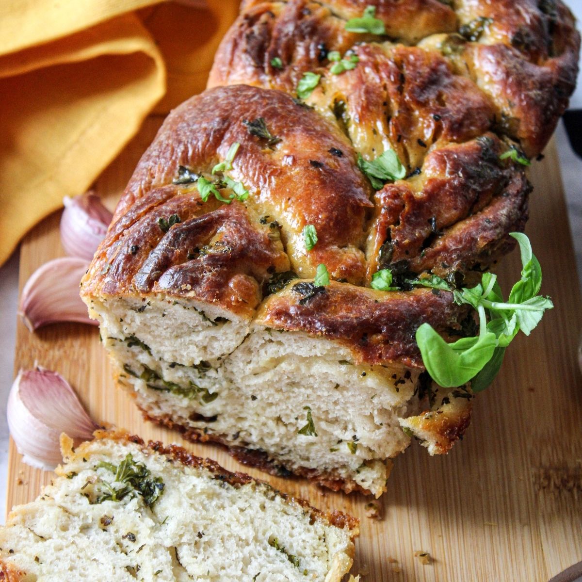 A sliced loaf of vegan garlic bread rests on a wooden board, garnished with fresh herbs, with garlic cloves and a yellow cloth in the background.