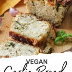 A sliced loaf of homemade garlic bread with herb swirls sits on a wooden board, garnished with fresh herbs and accompanied by garlic cloves.