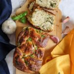 A loaf of homemade garlic bread with herb swirls is sliced on a wooden board, surrounded by fresh garlic, basil, and cloth serviettes—a perfect vegan garlic bread for any occasion.