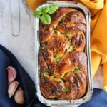 A loaf of homemade vegan garlic bread, plaited and garnished with basil, sits in a foil tray. Garlic cloves and a garlic bulb are nearby, with yellow and navy cloths adding colour in the background.