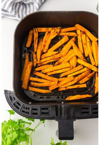 Frozen sweet potato chips are arranged in a single layer inside an air fryer basket, ready to be cooked or served. A striped tea towel and fresh herbs are nearby on a white surface.