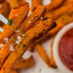 A fork holds three crispy air fryer sweet potato chips topped with coarse salt, with more frozen sweet potato chips and a bowl of tomato ketchup in the background. Text reads "Air Fryer Sweet Potatoes.