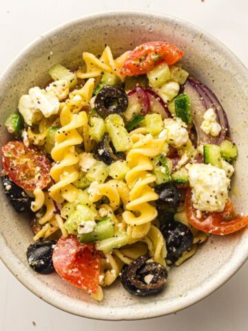 Bowl of pasta salad with spirali, cherry tomatoes, cucumber, black olives, red onion, and feta cheese, seasoned with herbs.
