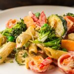 Pasta with mixed vegetables including broccoli, peas, courgette, and peppers, coated in a creamy sauce, served on a white plate.