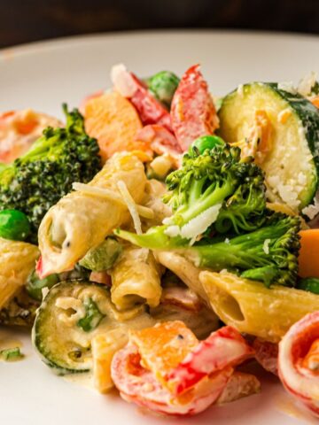 Pasta with mixed vegetables including broccoli, peas, courgette, and peppers, coated in a creamy sauce, served on a white plate.
