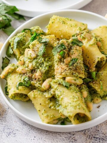 Plate of creamy pesto pasta with white beans and fresh herbs, garnished with ground pepper; uncooked pasta and basil leaves hint at this easy creamy pesto pasta recipe.