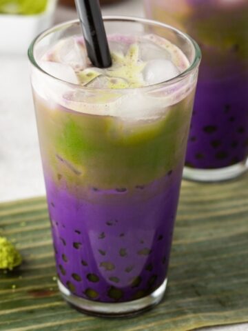 A glass of iced matcha milk bubble tea with ube creates striking purple and green layers, complemented by a black straw and boba pearls at the bottom, placed on a green mat.