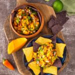 A bowl of mango salsa with diced vegetables, surrounded by yellow and blue corn tortilla crisps, sits on a wooden board next to a sliced mango, lime, and a yellow chilli pepper.