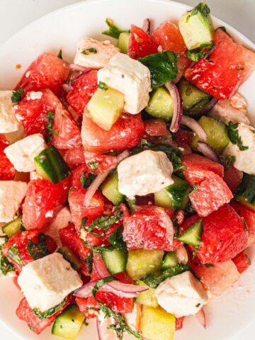 A salad with watermelon cubes, cucumber, feta cheese, red onion, and herbs on a white plate.