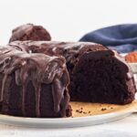 A sliced triple chocolate bundt cake with glossy chocolate icing drips, displayed on a white plate with a blue cloth in the background—an easy recipe perfect for chocolate lovers.