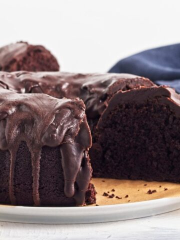 A sliced triple chocolate bundt cake with glossy chocolate icing drips, displayed on a white plate with a blue cloth in the background—an easy recipe perfect for chocolate lovers.