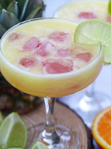 A yellow cocktail with ice and a lime wheel garnish in a coupe glass, with pineapple, lime wedges, and an orange in the background.