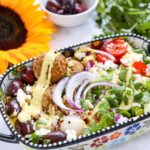A colourful salad with falafel, cherry tomatoes, red onion, kalamata olives, feta cheese, dressing, and greens in a decorative dish, with a sunflower and a bowl of olives nearby.
