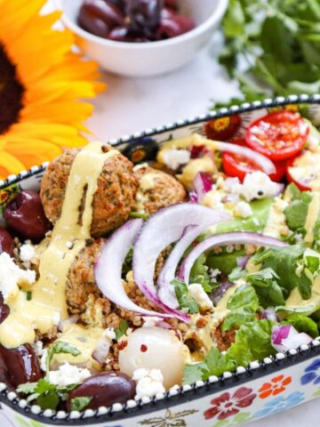 A colourful salad with falafel, cherry tomatoes, red onion, kalamata olives, feta cheese, dressing, and greens in a decorative dish, with a sunflower and a bowl of olives nearby.