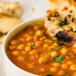 Close-up of a bowl of chickpea curry garnished with coriander, served with a piece of naan bread.