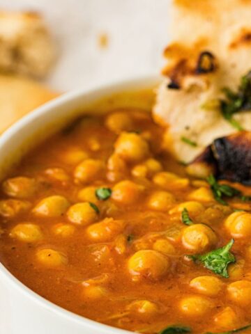 Close-up of a bowl of chickpea curry garnished with coriander, served with a piece of naan bread.