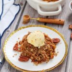 A portion of pecan crumble topped with a scoop of vanilla ice cream on a white plate with gold trim, garnished with whole pecans.