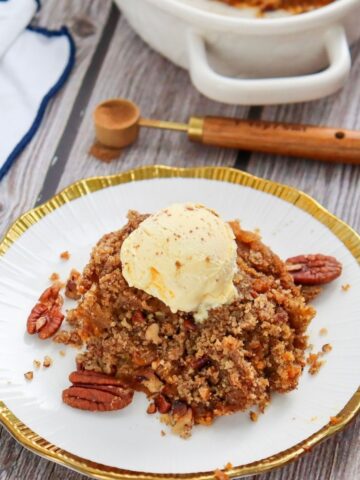 A portion of pecan crumble topped with a scoop of vanilla ice cream on a white plate with gold trim, garnished with whole pecans.