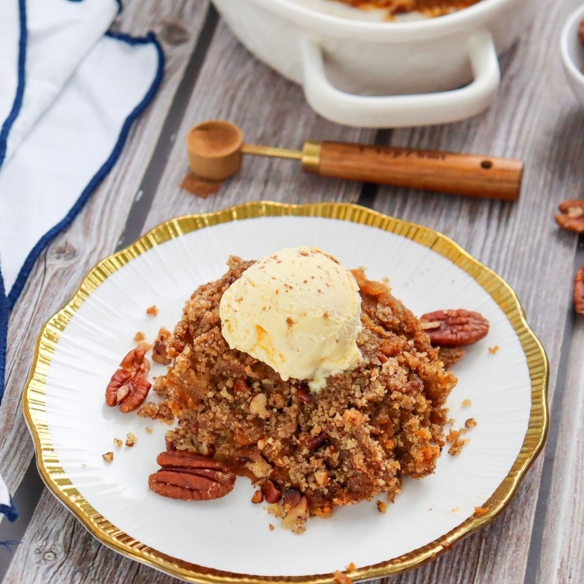 A portion of pecan crumble topped with a scoop of vanilla ice cream on a white plate with gold trim, garnished with whole pecans.