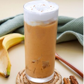 A tall glass of orange smoothie topped with whipped cream, beside a banana, cinnamon sticks, and a green cloth on a beige surface.
