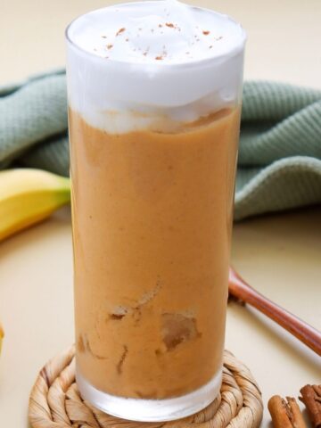 A tall glass of orange smoothie topped with whipped cream, beside a banana, cinnamon sticks, and a green cloth on a beige surface.
