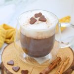 A glass mug of hot chocolate with frothy milk, topped with chocolate heart shapes, sits on a wooden coaster beside cinnamon sticks.