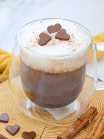 A glass mug of hot chocolate with frothy milk, topped with chocolate heart shapes, sits on a wooden coaster beside cinnamon sticks.