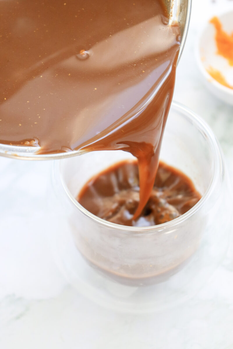 Pumpkin Spice Hot Chocolate Recipe (Vegan) 159 Chocolate sauce is being poured from a glass bowl into a clear glass tumbler on a white surface.