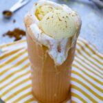 A tall glass filled with a creamy pumpkin spice drink topped with vanilla ice cream and cinnamon, sitting on a yellow striped cloth.