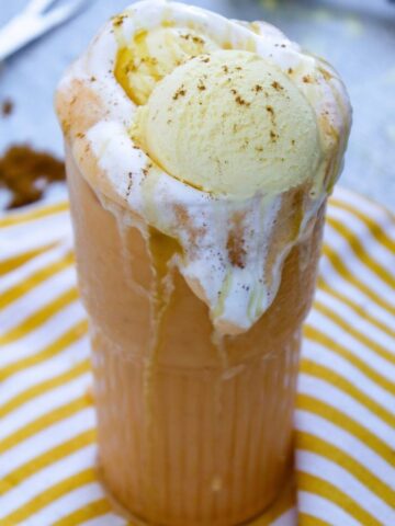 A tall glass filled with a creamy pumpkin spice drink topped with vanilla ice cream and cinnamon, sitting on a yellow striped cloth.