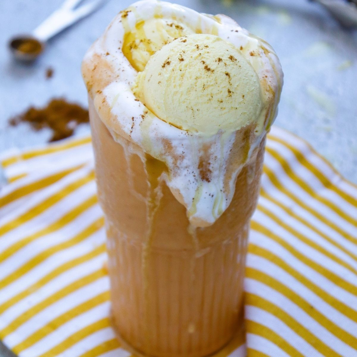 A tall glass filled with a creamy pumpkin spice drink topped with vanilla ice cream and cinnamon, sitting on a yellow striped cloth.