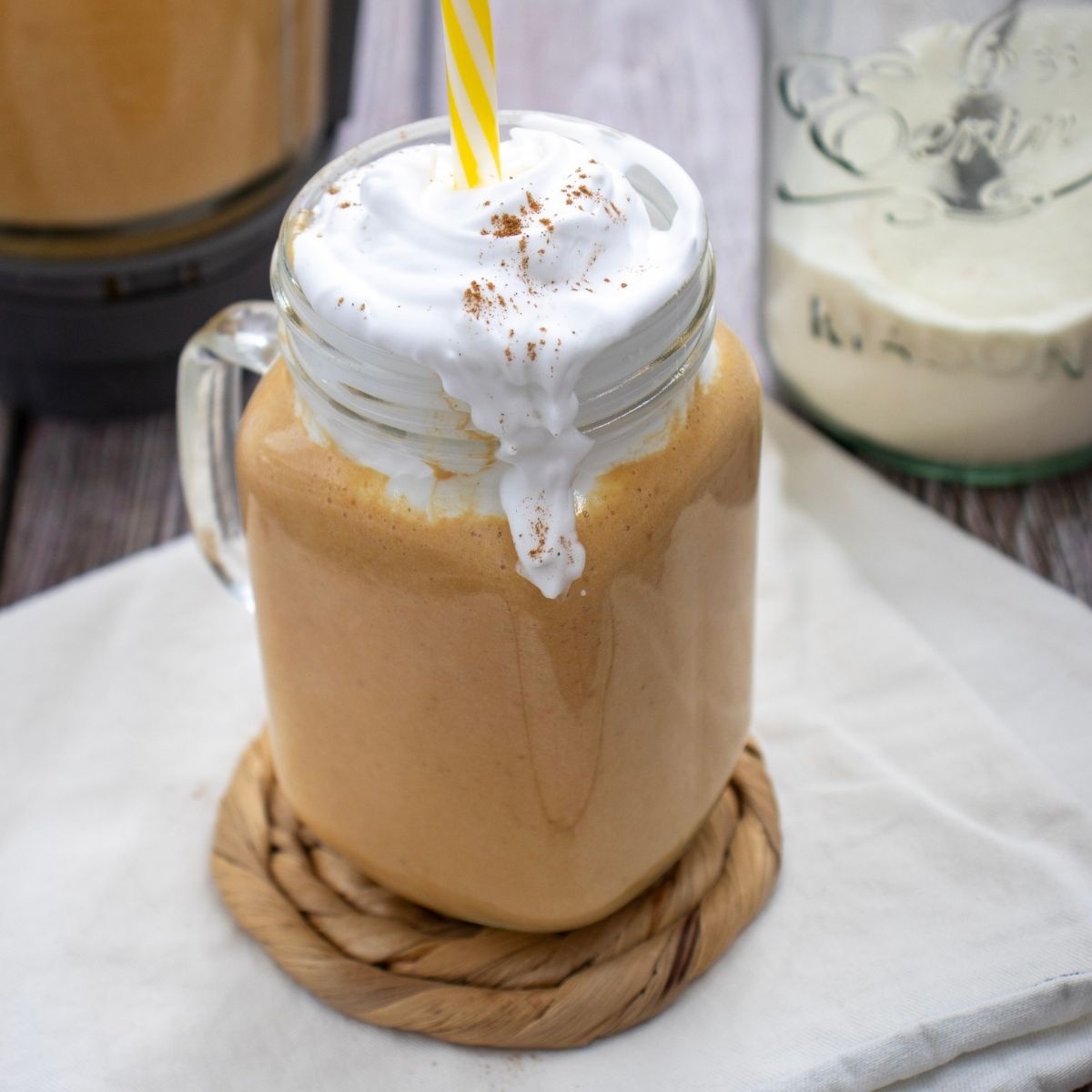 A jam jar filled with a light brown smoothie, topped with whipped cream and a yellow straw.
