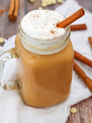 A jam jar filled with a beige drink, topped with whipped cream and a cinnamon stick.